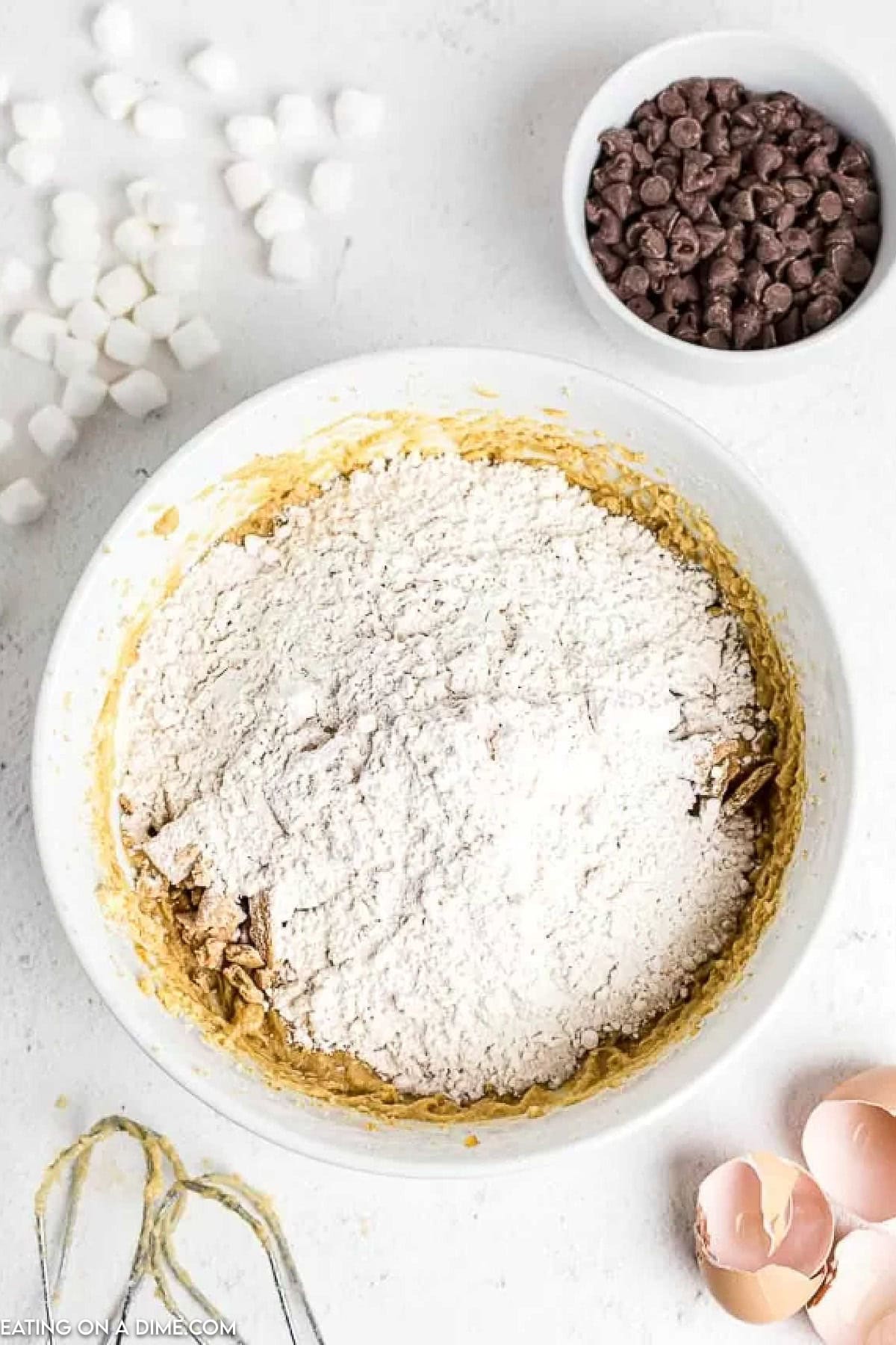 A white bowl filled with flour, chopped nuts, and dough mixture rests on a white surface. Nearby, essential ingredients for s’mores cookies await: a bowl of chocolate chips, scattered marshmallows, a cracked egg with shells, and a whisk attachment.