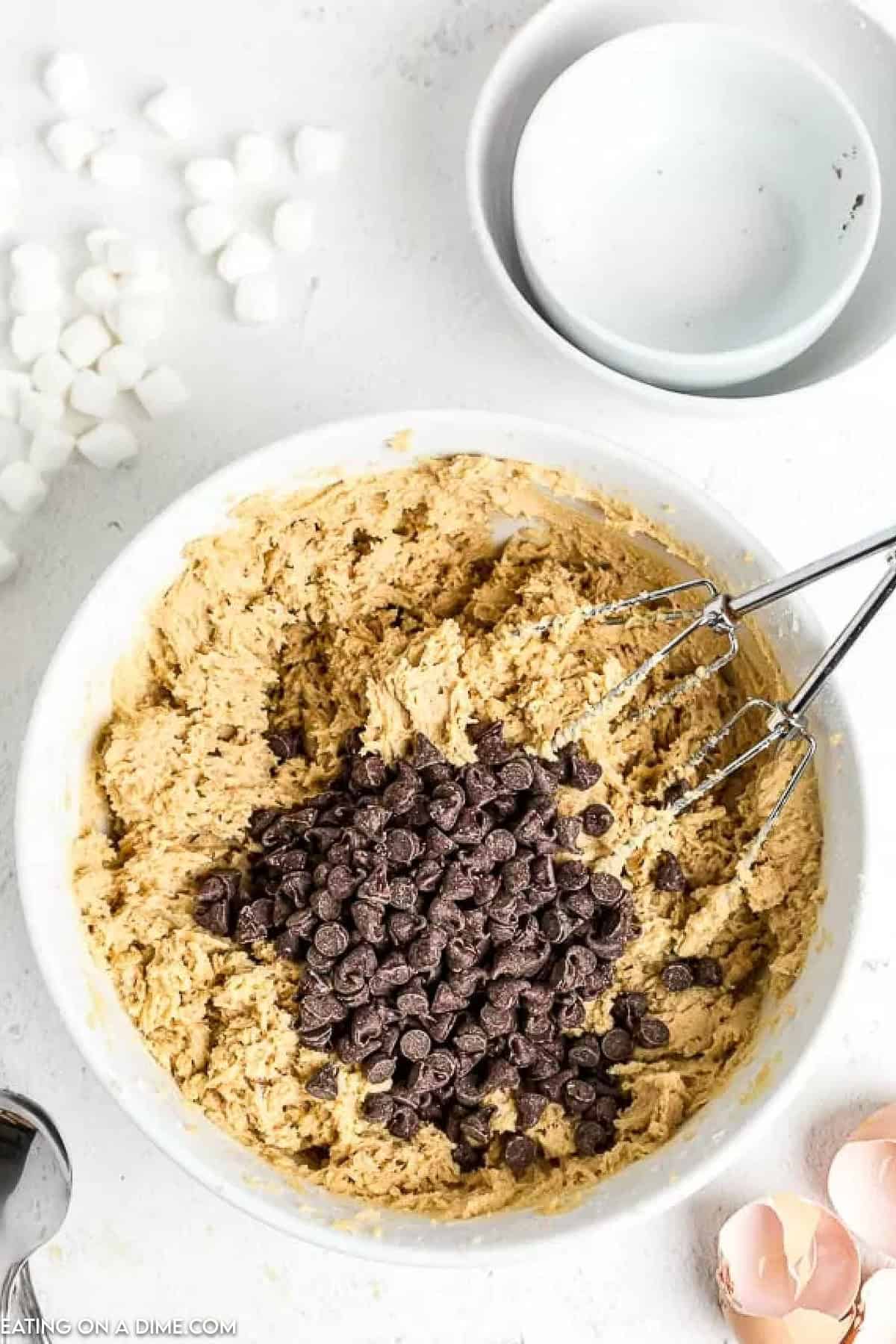 A mixing bowl brimming with chocolate chip cookie dough and chocolate chips is surrounded by a whisk, empty bowls, eggshells, and marshmallows on a pristine white surface—hinting at the creation of irresistible s'mores cookies.