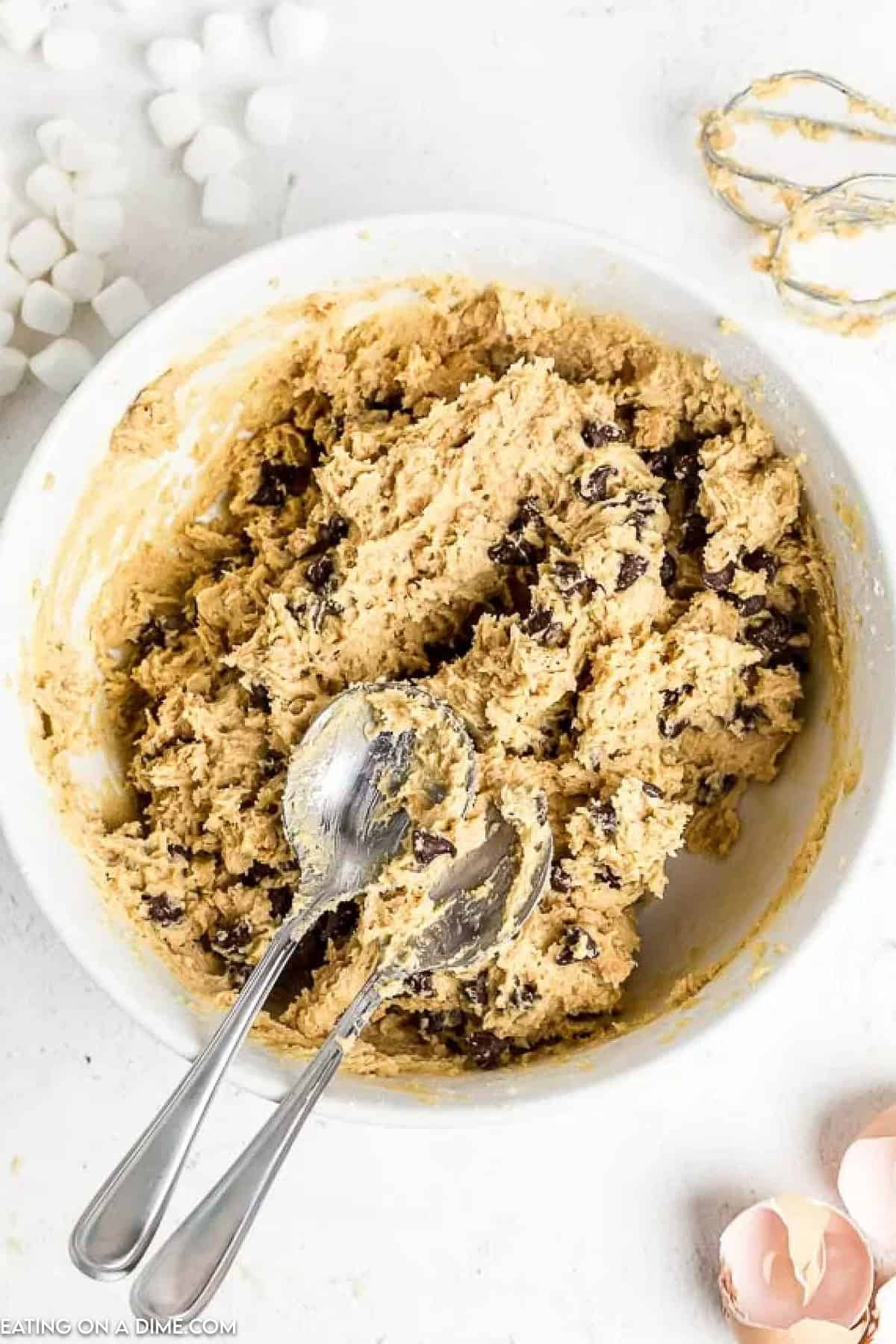 A bowl filled with chocolate chip cookie dough sits on a white surface, ready to become S'mores Cookies. Two metal spoons rest in the bowl, surrounded by broken eggshells and marshmallows. A whisk is casually placed at the top right corner, hinting at the sweet creation to come.