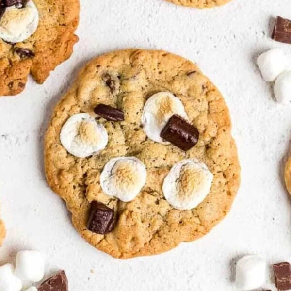 A scrumptious s'mores cookie with marshmallows and chocolate chunks rests on a pristine white surface. Surrounding it are pieces of chocolate and small marshmallows, with other cookies peeking in from the edges.