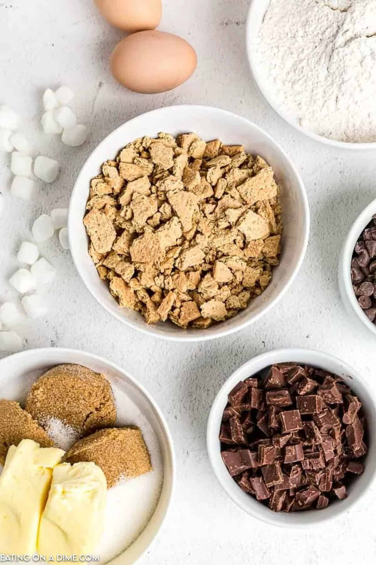 Bowls of baking ingredients for S'mores Cookies are spread across the countertop, featuring crushed graham crackers, flour, chocolate chips and chunks, alongside two eggs, marshmallows, and a bowl with butter and sugar.