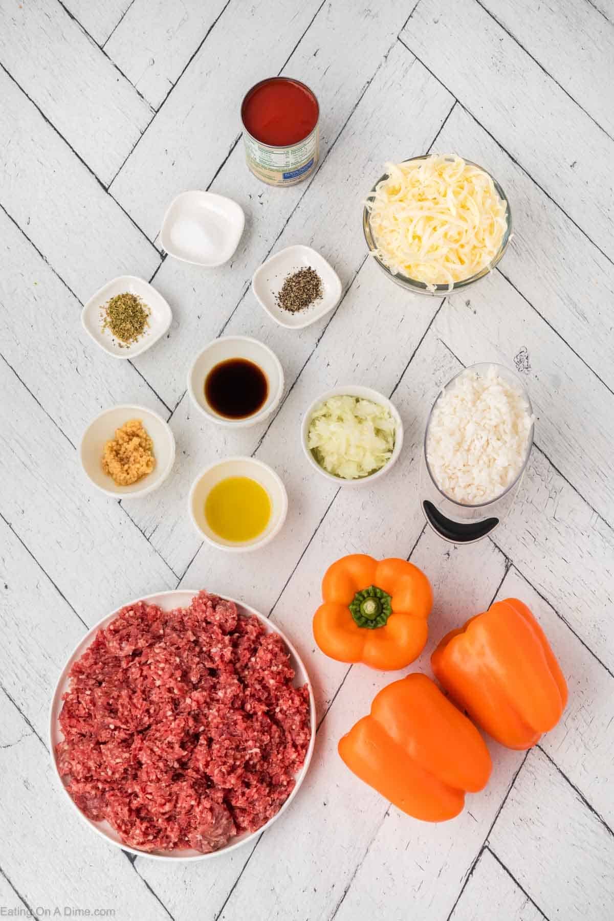 A recipe for stuffed peppers unfolds on a white wooden surface: ground beef, two orange bell peppers, minced garlic in a small bowl, olive oil, chopped onions, shredded cheese, a can of tomato sauce, and spices neatly arranged in small dishes. Perfect for crafting the ideal dish.