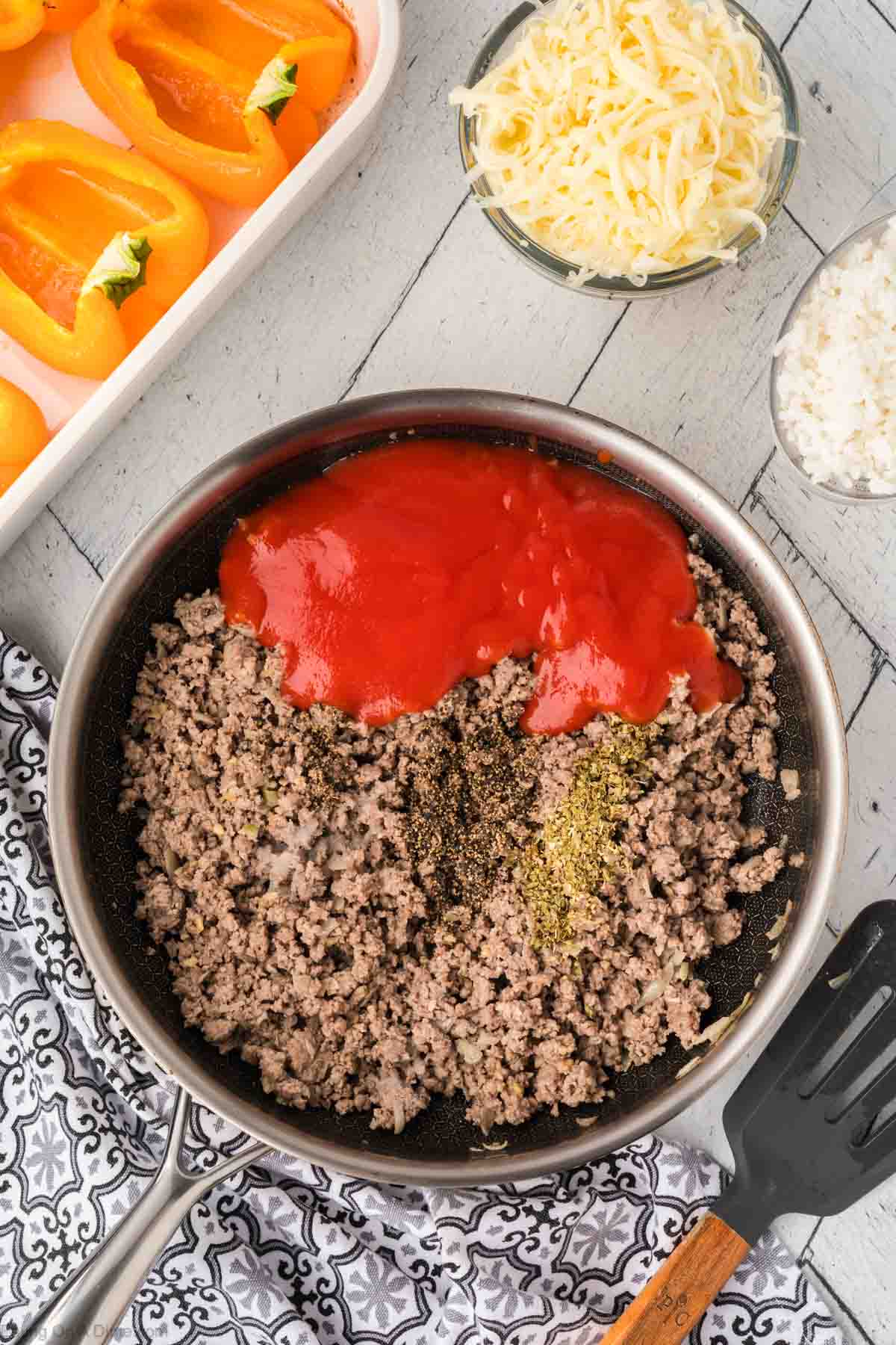 A skillet on a patterned cloth holds ground meat mixed with tomato sauce and spices, perfect for stuffed peppers. Beside it are halved bell peppers in a baking dish, ready to be filled, alongside a bowl of grated cheese and chopped onions on a wooden surface.