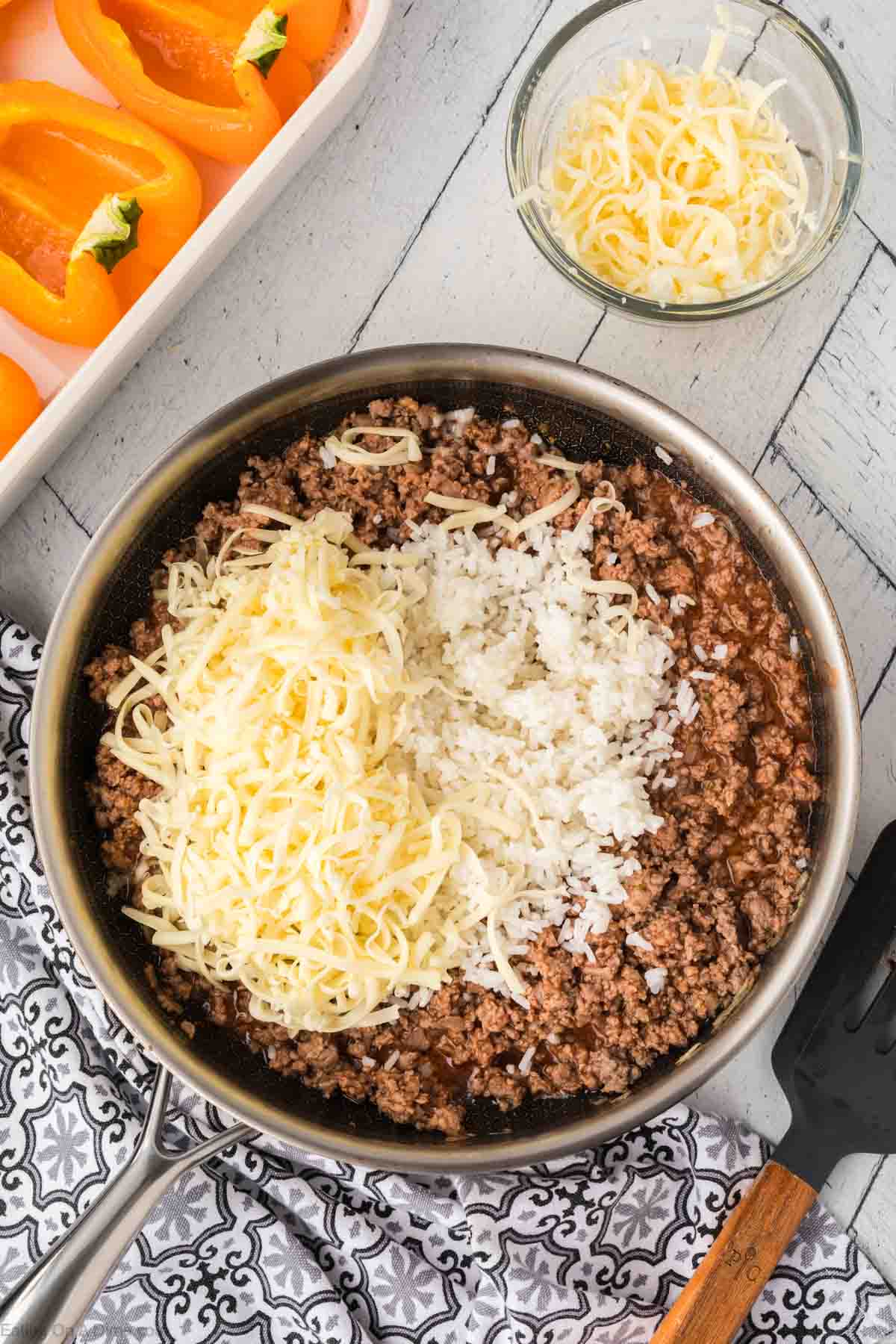 A skillet filled with cooked ground beef topped with shredded cheese and chopped onions sits alongside a white tray showcasing sliced orange bell peppers, perfect for making stuffed peppers. Nearby, a small glass bowl holds extra cheese, and a black spatula completes the scene.