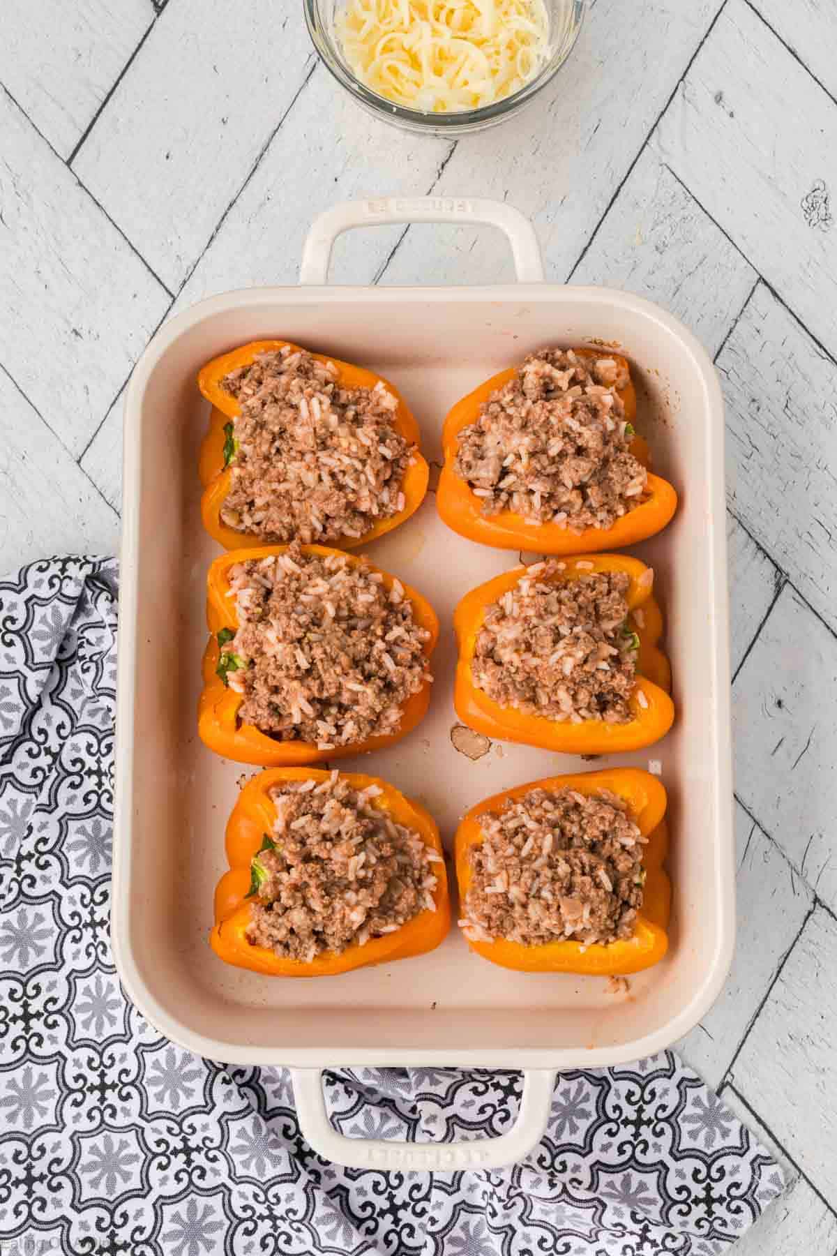 A white baking dish holds six orange bell pepper halves, each artfully stuffed with a savory rice and ground meat blend. Resting on a light wooden surface, the scene is completed with a peek of a patterned cloth and a bowl of shredded cheese in the background.