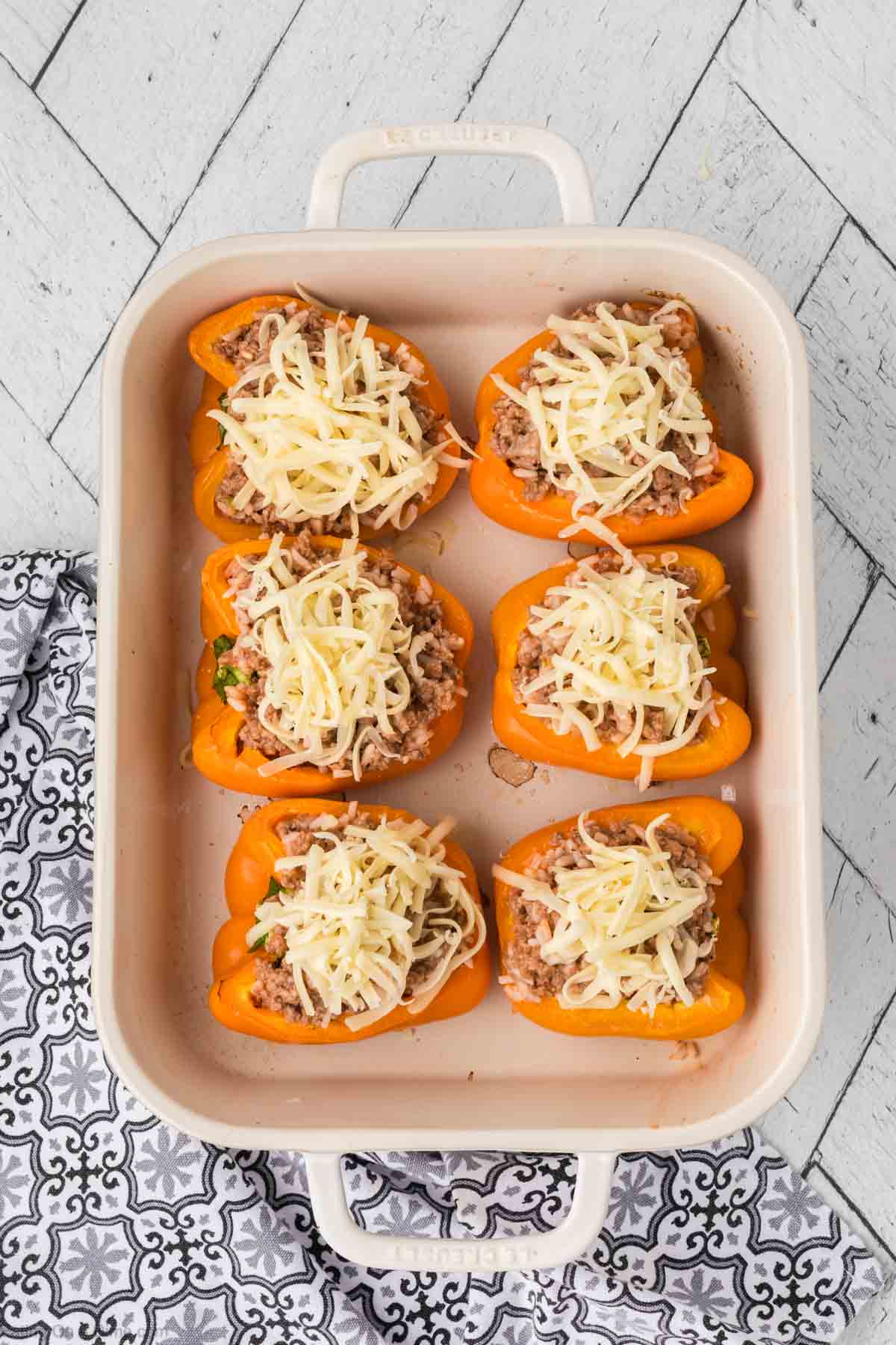 A baking dish holds six stuffed peppers, each orange bell pepper half brimming with a savory blend of minced meat and herbs, crowned with shredded cheese. The dish rests on a patterned towel atop a white, textured surface, ready for the oven.