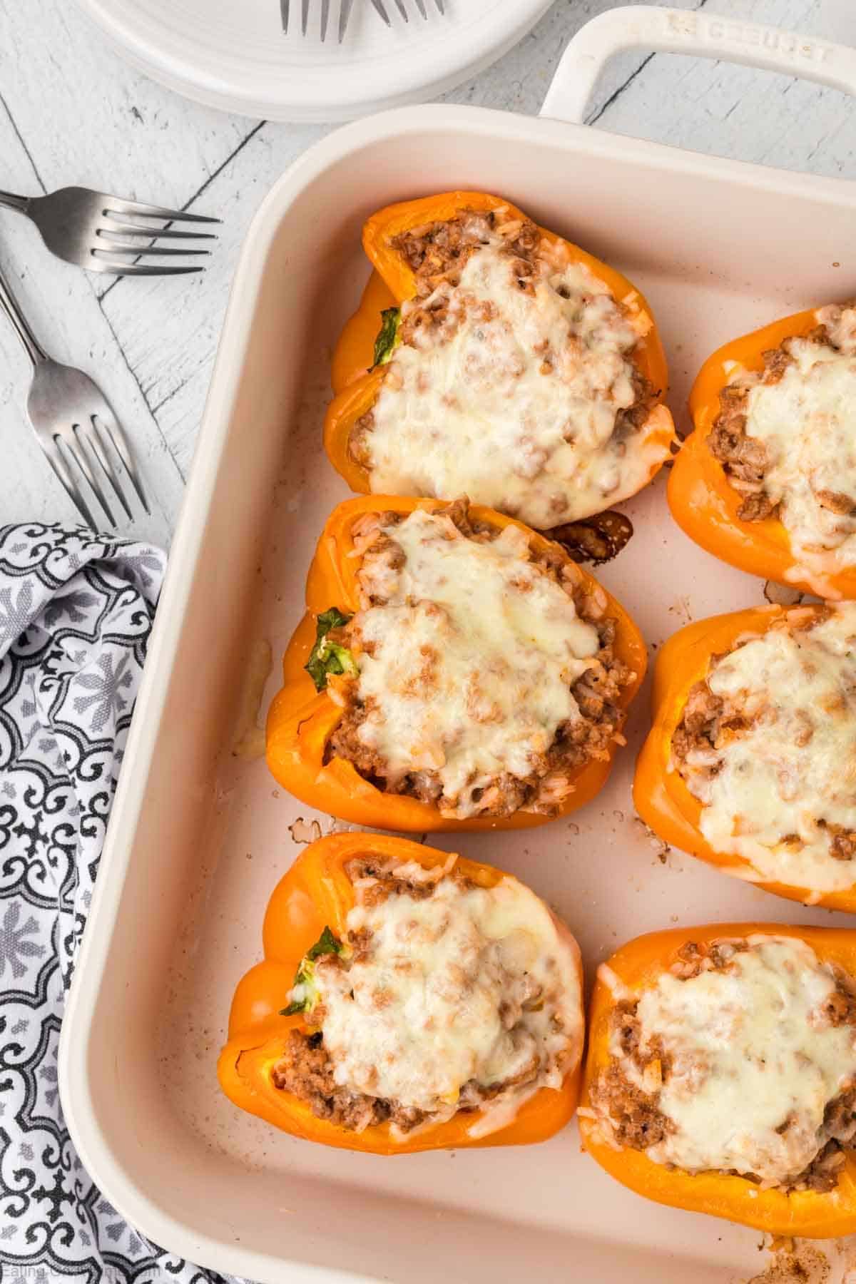 A baking dish showcases six stuffed orange bell peppers topped with melted cheese, embodying a delightful recipe. Each pepper is neatly arranged and filled with a savory mixture. Nearby are three forks and a patterned napkin on a white wooden surface, ready for an enjoyable cooking experience.