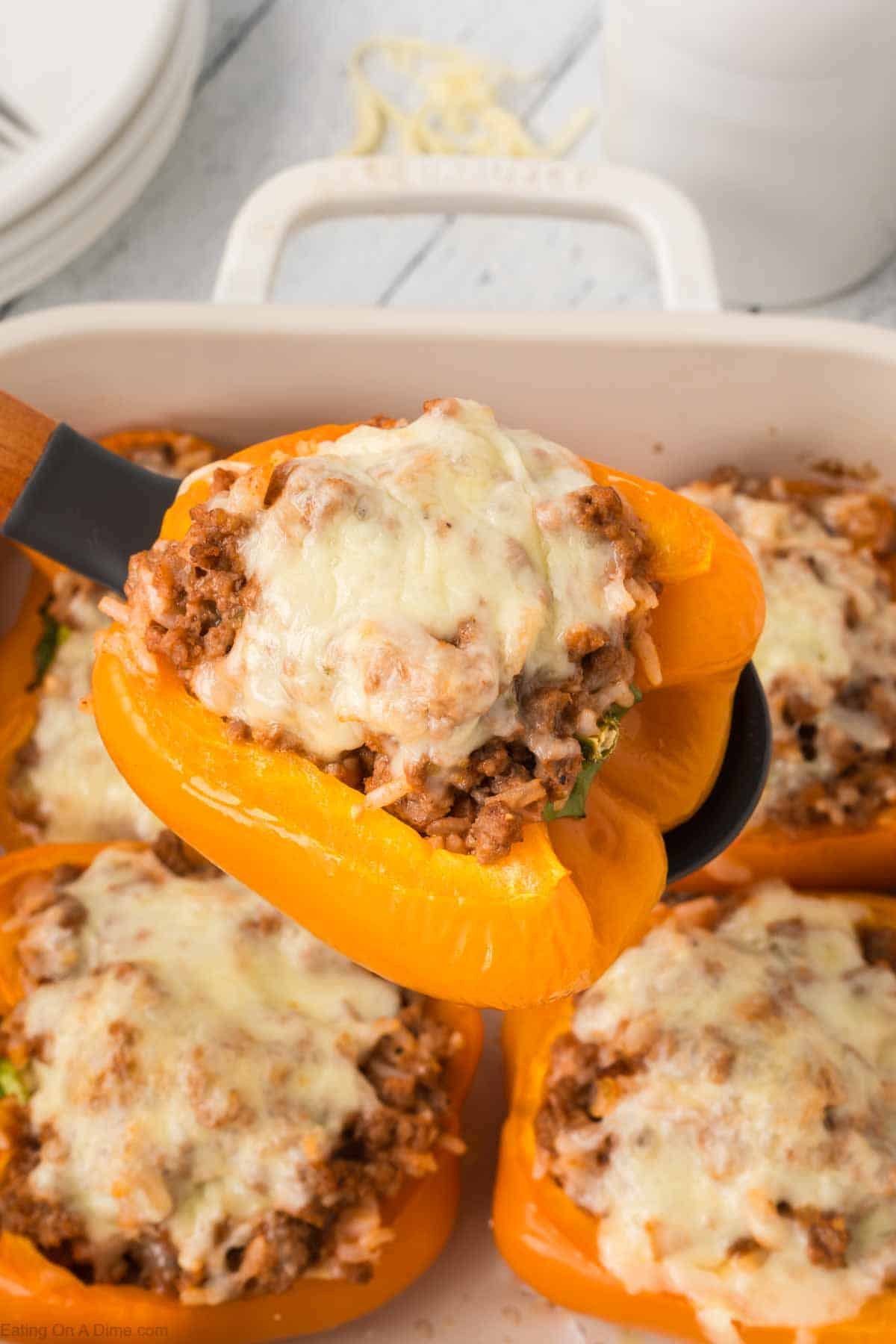 A baking dish holds delicious Stuffed Peppers, orange bell beauties topped with melted cheese and filled with a savory mix of ground meat and rice. One pepper is being lifted with a spatula, oozing cheesy goodness.