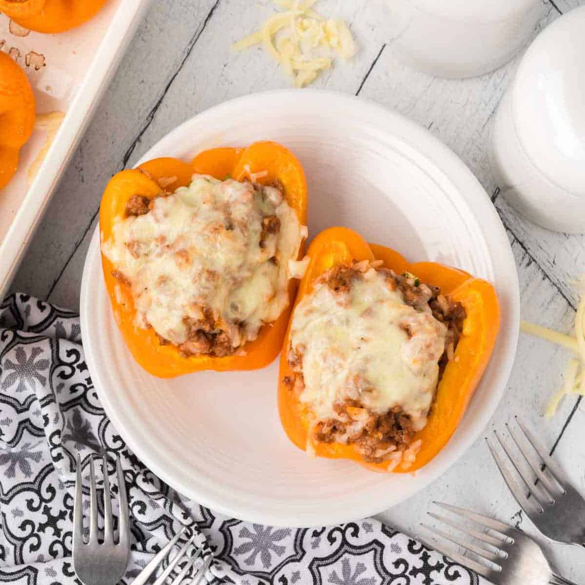 Two orange bell peppers brimming with a savory meat and rice mixture, topped with melted cheese, sit on a white plate on a rustic wooden table. Nearby, a patterned napkin and forks await the delicious stuffed peppers.