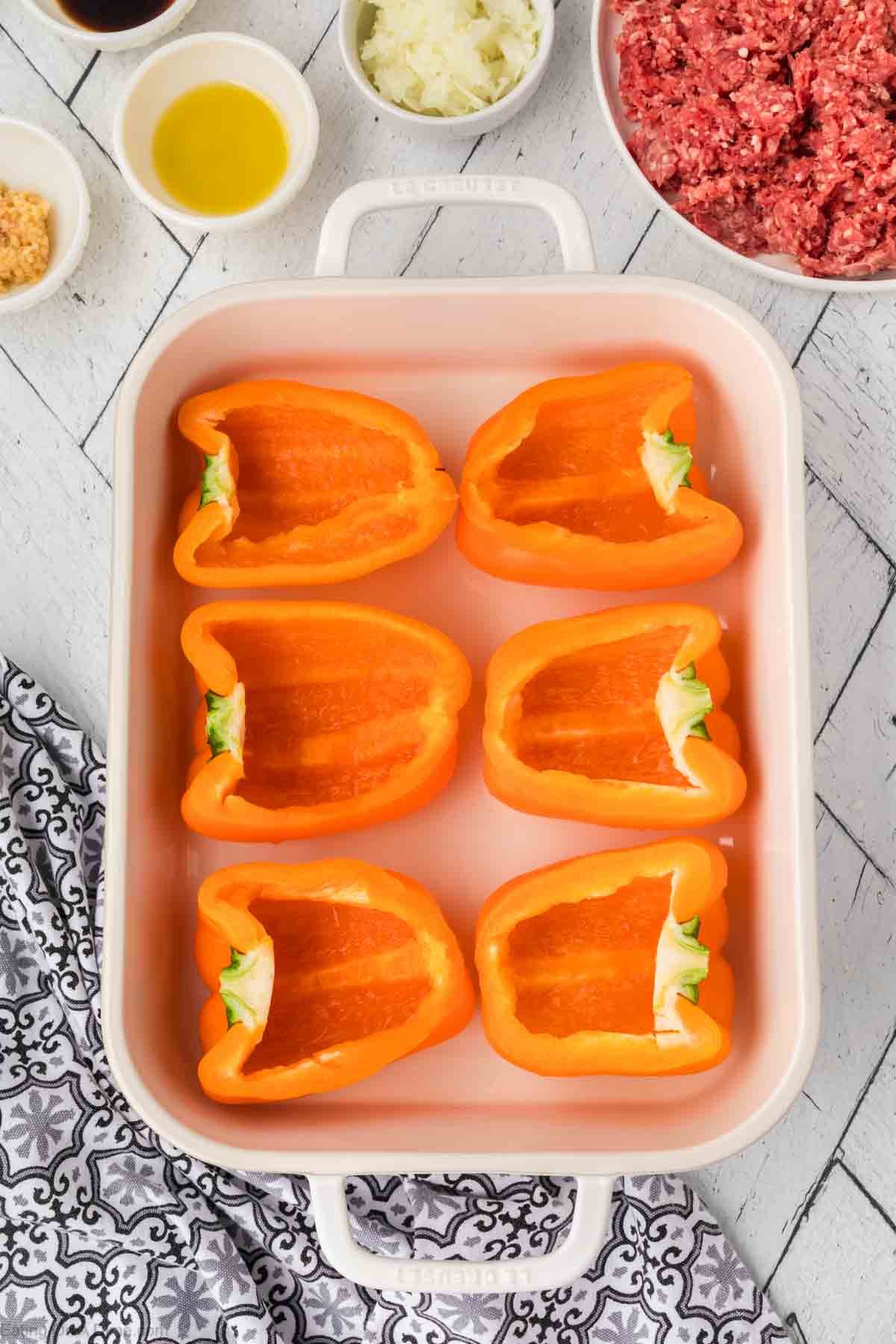 Orange bell pepper halves are poised for transformation into stuffed peppers in a white rectangular baking dish. Surrounding them, small bowls cradle minced onions, ground meat, oil, spices, and sauces. The setup rests on a patterned surface with a black and white cloth beside it.