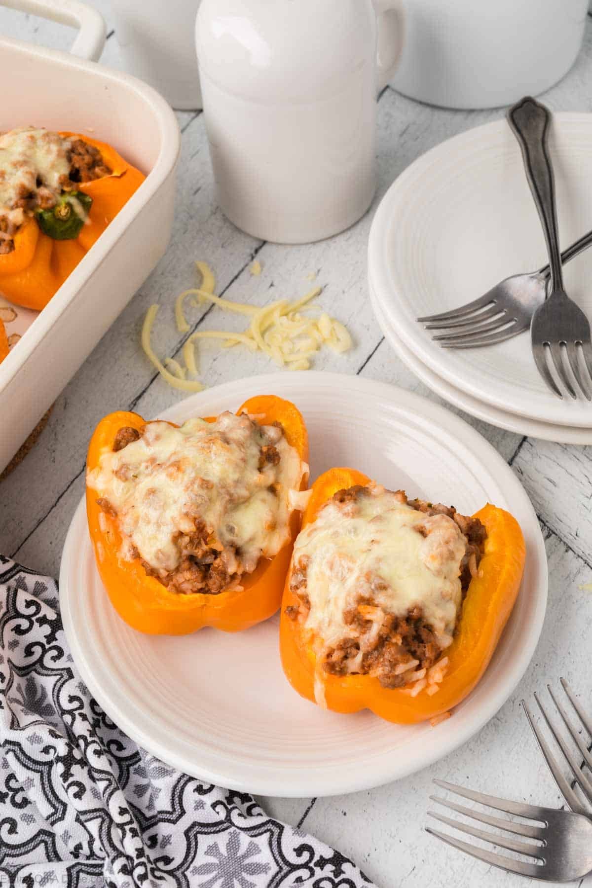 Two stuffed orange bell peppers, brimming with minced meat and topped with melted cheese, rest invitingly on a white plate. Nearby are forks, additional white plates, and a black and white patterned napkin on a light wooden table—perfect accompaniments to these delectable stuffed peppers.