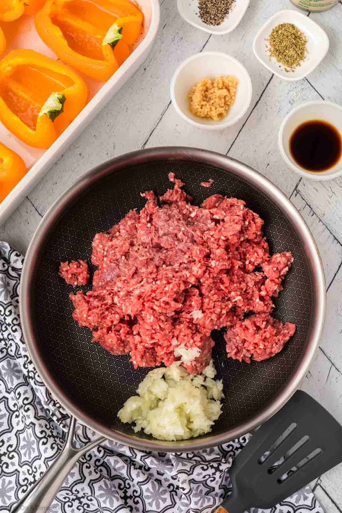 A frying pan with raw ground beef and chopped onions sits ready for a tasty dish. Nearby, yellow bell pepper halves await stuffing with herbs, minced garlic, and a touch of soy sauce. A black and white patterned cloth and a spatula complete this stuffed peppers preparation scene.