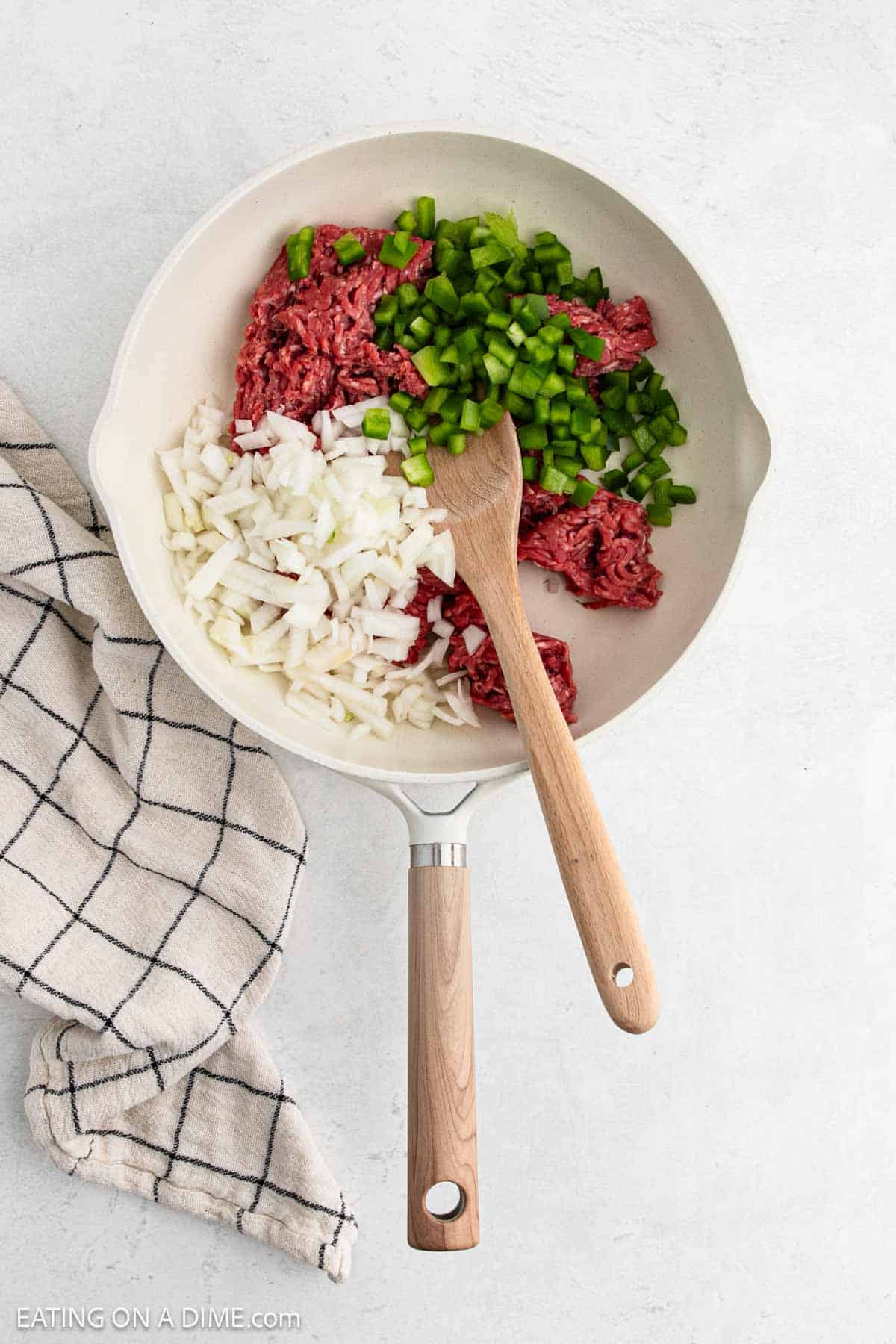 Cooking ground beef in a large skillet with diced green bell peppers and onions with a wooden spoon