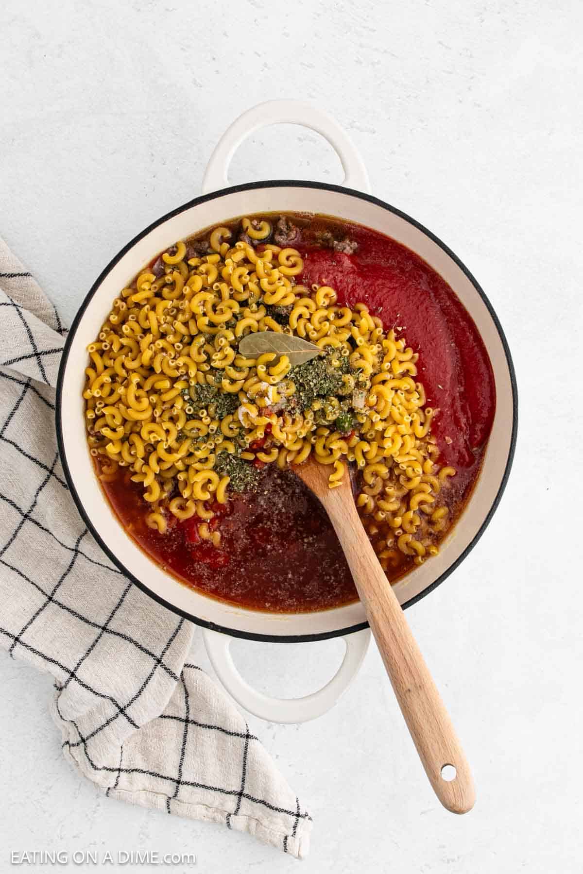 Elbow macaroni noodles, tomato sauce, and seasoning in a large pot with a wooden spoon